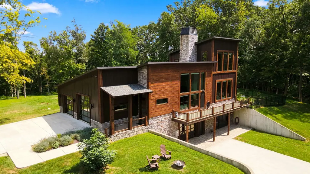 Aerial view of the house in full summer canopy, with the stone chimney, glass garage doors, deck, walkout lower level, and fenced yard.